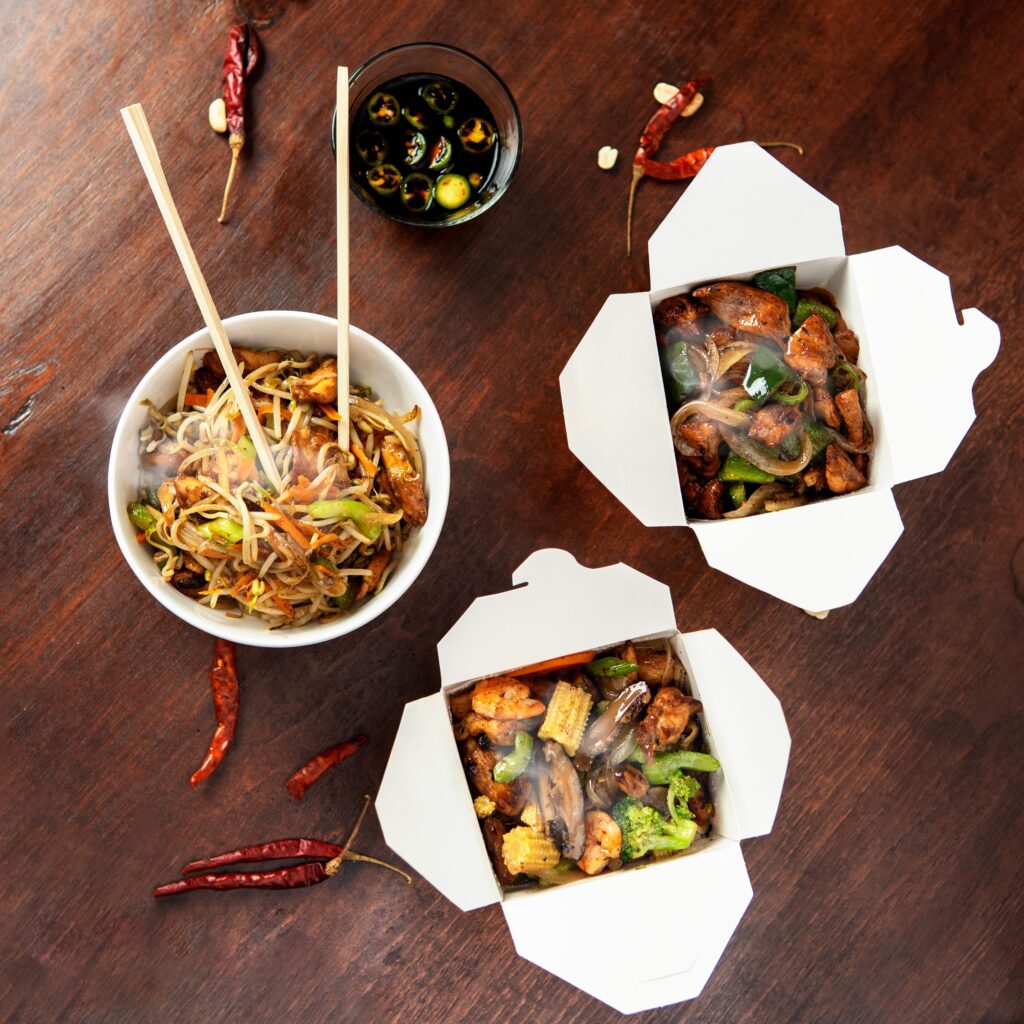 Flat lay of Chinese cuisine with noodles and vegetables in takeout boxes, served with chopsticks.